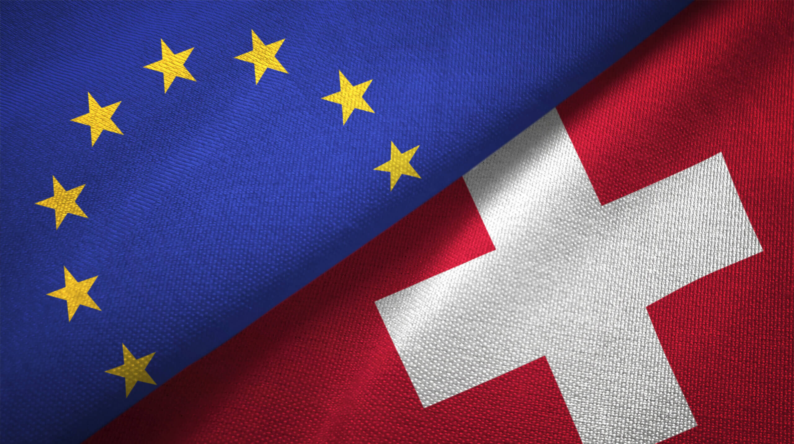 Das Bild zeigt die Flaggen der Europäischen Union (EU) und der Schweiz, die sich diagonal berühren. Die EU-Flagge, erkennbar durch ihren blauen Hintergrund und den Kreis aus 12 goldenen Sternen, liegt oben links im Bild. Diese Symbole repräsentieren Einheit, Solidarität und Frieden in Europa. Entgegengesetzt dazu befindet sich unten rechts im Bild die Flagge der Schweiz. Ihr roter Hintergrund und das weiße Kreuz stehen für die Geschichte, Tradition und Neutralität des Landes. Die diagonalen Positionen der Flaggen erzeugen eine visuelle Verbindung und symbolisieren möglicherweise die engen politischen und wirtschaftlichen Beziehungen zwischen der EU und der Schweiz. Die Flaggen füllen das gesamte Bild aus, was ihre dominante Präsenz und Bedeutung unterstreicht. Die klare und kraftvolle Darstellung der Flaggen betont die Wichtigkeit der beiden politischen Einheiten in Europa. Insgesamt strahlt das Bild eine Atmosphäre von Stabilität, Zusammenarbeit und Respekt aus. Es lädt den Betrachter dazu ein, über die Beziehungen zwischen verschiedenen Ländern und politischen Einheiten nachzudenken und die Vielfalt und Einheit Europas zu würdigen.